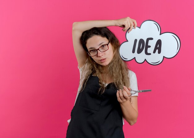 Jeune femme de coiffeur professionnel en tablier tenant des ciseaux et signe de bulle de discours avec idée de mot souriant confiant