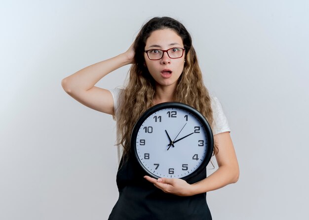 Jeune femme de coiffeur professionnel en tablier tenant horloge murale regardant à l'avant confus debout sur un mur blanc