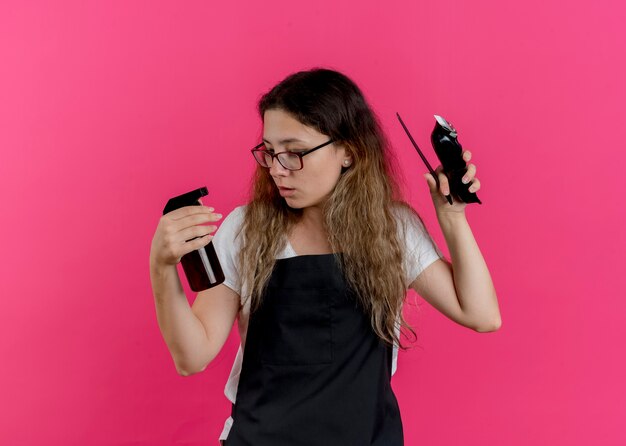 Jeune femme de coiffeur professionnel en tablier tenant un peigne tondeuse et un spray à la confusion essayant de faire un choix