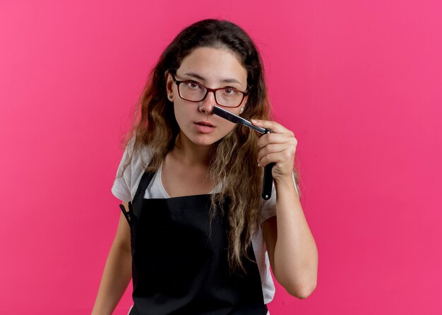 Jeune femme de coiffeur professionnel en tablier tenant un rasoir avec une expression sérieuse