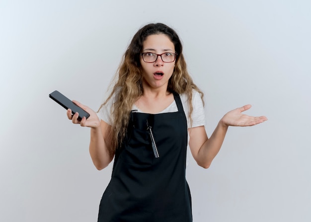 Jeune femme de coiffeur professionnel en tablier tenant le smartphone à l'avant en haussant les épaules d'être confus et mécontent de se tenir debout sur un mur blanc