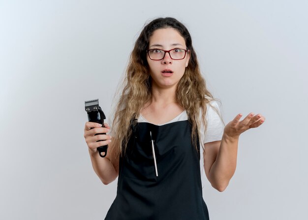 Jeune femme de coiffeur professionnel en tablier tenant tondeuse à l'avant avec le bras d'être surpris et confus debout sur un mur blanc