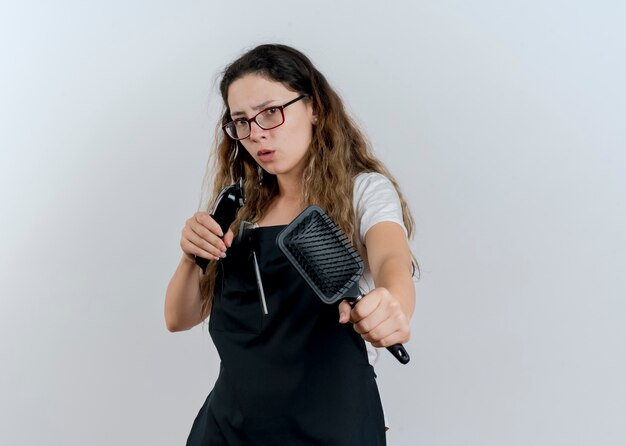 Jeune femme de coiffeur professionnel en tablier tenant une tondeuse et une brosse à cheveux à l'avant d'être inquiet debout sur un mur blanc