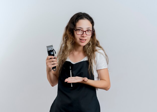 Jeune femme de coiffeur professionnel en tablier tenant tondeuse le présentant avec le bras de sa main souriant debout sur un mur blanc