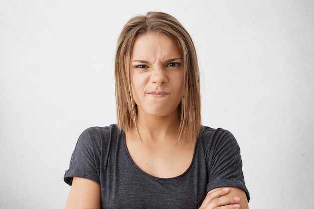 Jeune femme en colère aux yeux noirs fronçant le nez et les lèvres portant un T-shirt décontracté debout mains croisées isolées montrant son insatisfaction. Concept de personnes et d'émotions