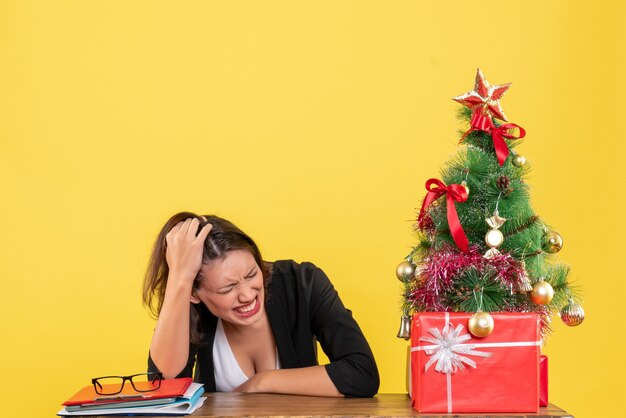 Jeune femme en colère en costume près de sapin de Noël décoré au bureau sur jaune