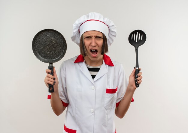 Jeune femme en colère cuisinier en uniforme de chef tenant une spatule et une poêle à la recherche d'isolement sur le blanc