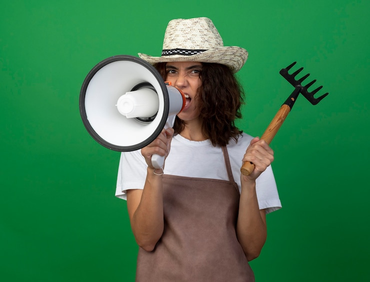 Photo gratuit jeune femme en colère jardinier en uniforme portant chapeau de jardinage tenant râteau parle sur haut-parleur