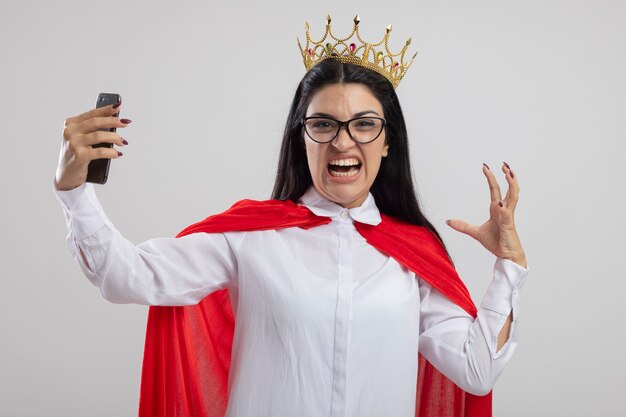 Jeune femme en colère portant des lunettes et une couronne tenant un téléphone mobile en gardant la main dans l'air à l'avant isolé sur un mur blanc