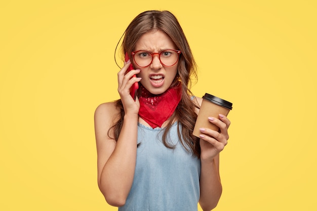 Une Jeune Femme En Colère Se Sent Intense Lorsqu'elle A Une Conversation Téléphonique Avec Un Ami, Entend Des Bêtises, N'est Pas D'accord Avec Quelque Chose, Sourit Avec Aversion, Boit Du Café, Isolée Sur Un Mur Jaune.