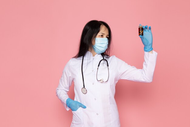 Jeune femme en combinaison médicale blanche gants bleus masque de protection bleu tenant des pilules colorées sur rose