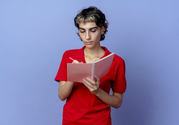 Jeune femme concentrée avec coupe de cheveux pixie écrit quelque chose sur le bloc-notes avec un stylo