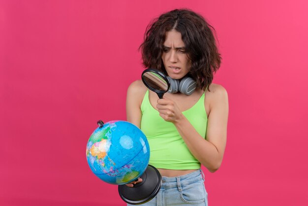Une jeune femme confuse aux cheveux courts en vert crop top dans les écouteurs regardant globe avec loupe