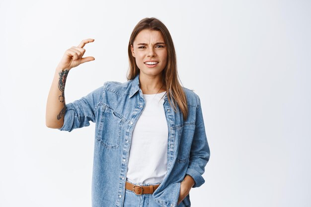 Jeune femme confuse et déçue, fronçant les sourcils, montrant une petite taille avec les doigts et regardant fixement. Fille se plaignant de petite chose, trop petite, debout sur blanc