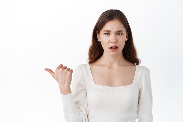 Jeune femme confuse pointant de côté une étrange bannière, regarde devant elle interrogée, posant une question, debout contre un mur blanc en studio