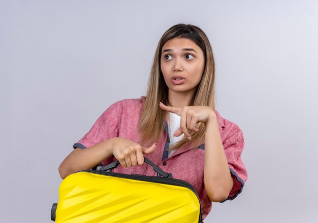 Une jeune femme confuse portant une chemise rouge tenant une valise jaune tout en pointant sur le côté avec l'index sur un mur blanc