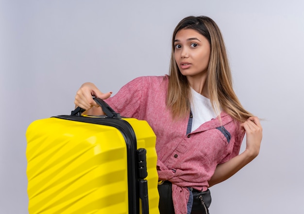 Une jeune femme confuse portant une chemise rouge tenant une valise jaune tout en regardant sur un mur blanc