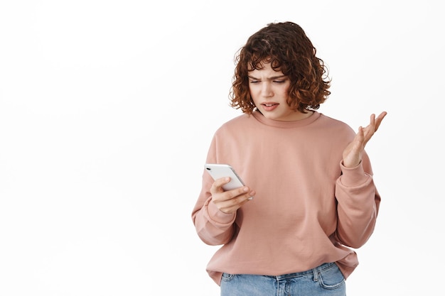Jeune femme confuse regardant l'écran du smartphone avec un visage agacé, ne peut pas comprendre ce qui se passe, fixe un message absurde sur le téléphone, debout sur fond blanc