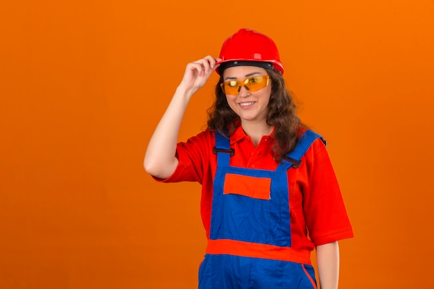 Jeune femme constructeur en construction lunettes uniformes et casque de sécurité avec sourire amical touchant le casque sur mur orange isolé