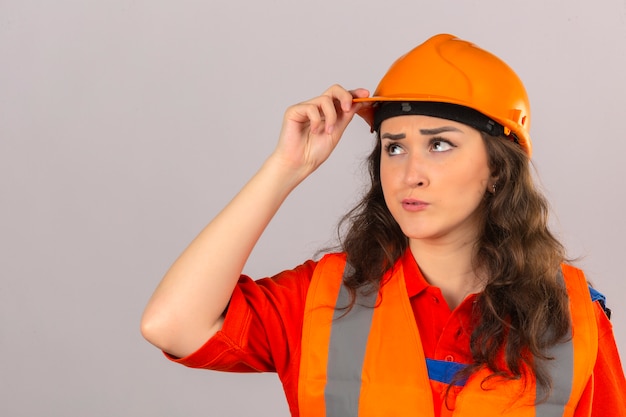 Jeune femme constructeur mécontent en uniforme de construction et casque de sécurité en levant et en pensant toucher son casque sur fond blanc isolé