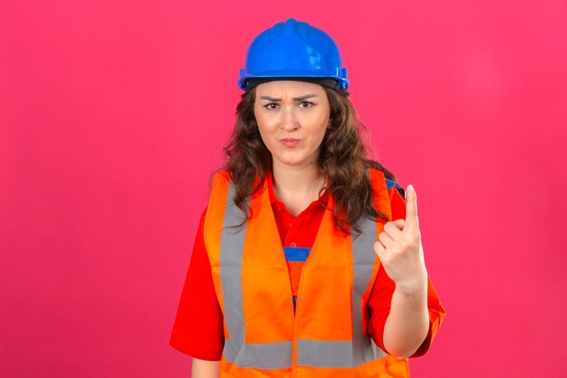 Jeune femme constructeur mécontent en uniforme de construction et casque de sécurité pointant vers le haut avec l'index d'avertissement sur fond rose