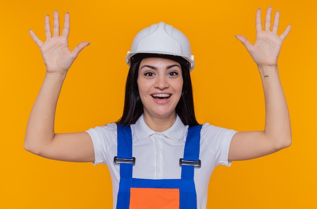 Jeune femme constructeur en uniforme de construction et casque de sécurité à l'avant souriant joyeusement heureux et positif avec les bras levés debout sur le mur orange