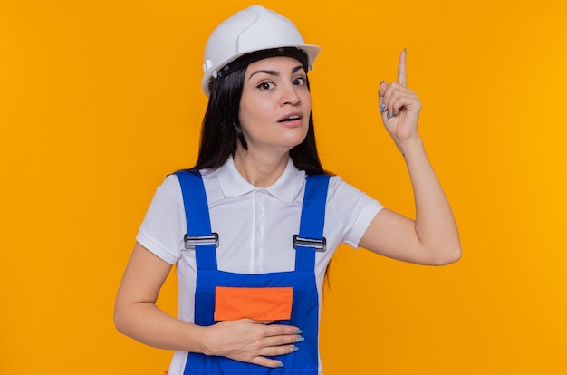 Jeune femme constructeur en uniforme de construction et casque de sécurité à l'avant avec le sourire sur le visage surpris intelligent montrant l'index ayant une excellente idée debout sur un mur orange