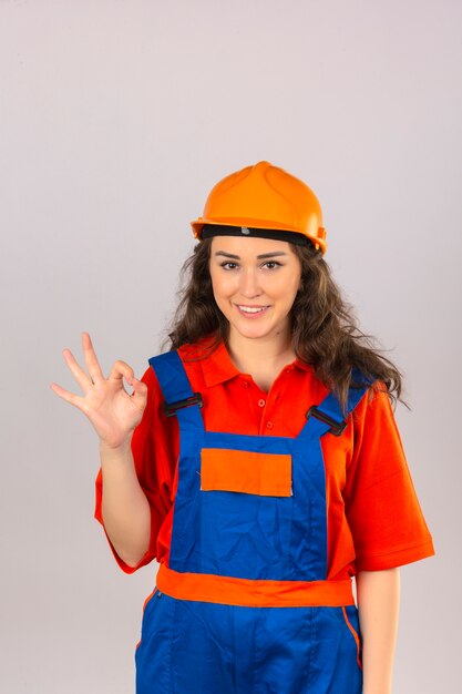 Jeune femme constructeur en uniforme de construction et casque de sécurité à la confiance en souriant joyeusement faisant signe ok sur mur blanc isolé