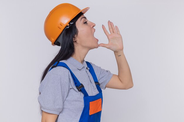 Jeune femme constructeur en uniforme de construction et casque de sécurité en criant ou en appelant quelqu'un hlding main sur la bouche debout sur un mur blanc