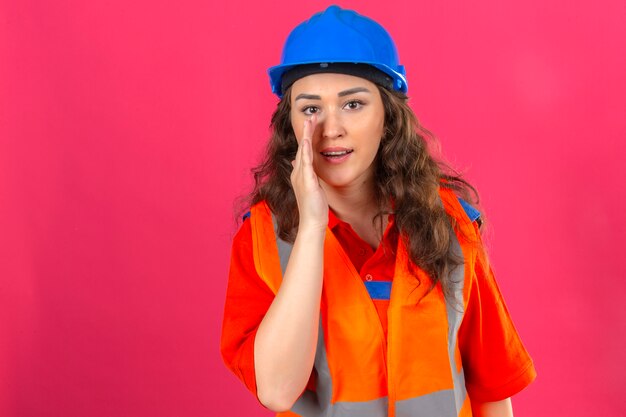 Jeune femme constructeur en uniforme de construction et casque de sécurité criant avec la main sur la bouche sur mur rose isolé