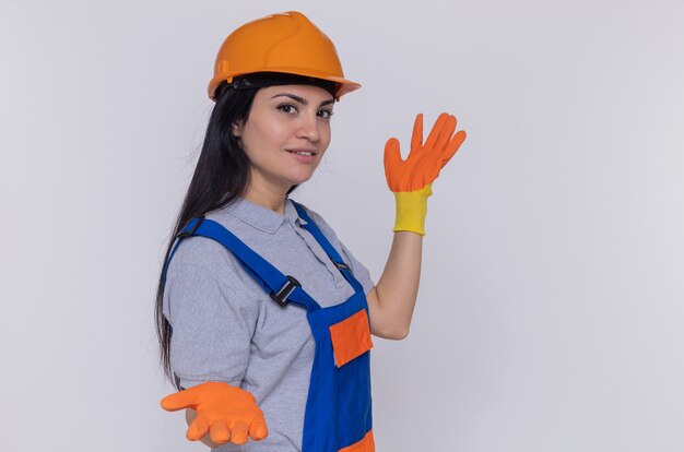 Jeune femme constructeur en uniforme de construction et casque de sécurité dans des gants en caoutchouc à l'avant souriant confiant présentant avec les bras debout sur un mur blanc