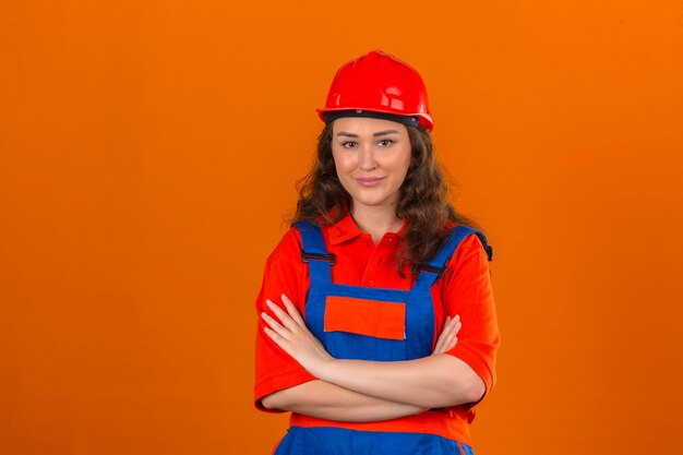 Jeune femme constructeur en uniforme de construction et casque de sécurité debout avec les bras croisés avec sourire sur le visage sur mur orange isolé