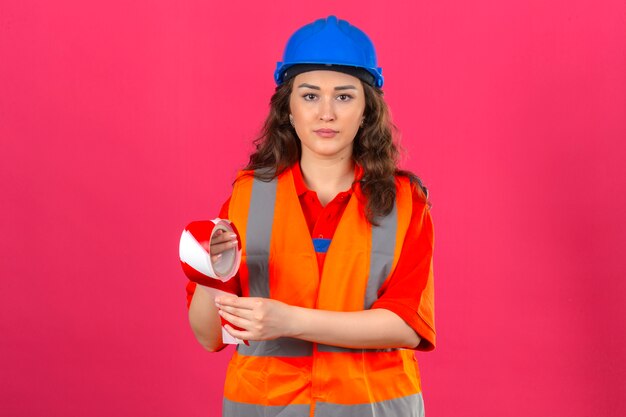 Jeune femme constructeur en uniforme de construction et casque de sécurité debout avec du ruban adhésif avec visage sérieux sur mur rose isolé_