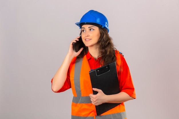 Jeune femme constructeur en uniforme de construction et casque de sécurité debout avec presse-papiers parler sur téléphone mobile sur mur blanc isolé