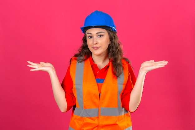 Photo gratuite jeune femme constructeur en uniforme de construction et casque de sécurité désemparé et expression confuse avec les bras et les mains soulevées concept de doute sur mur rose isolé