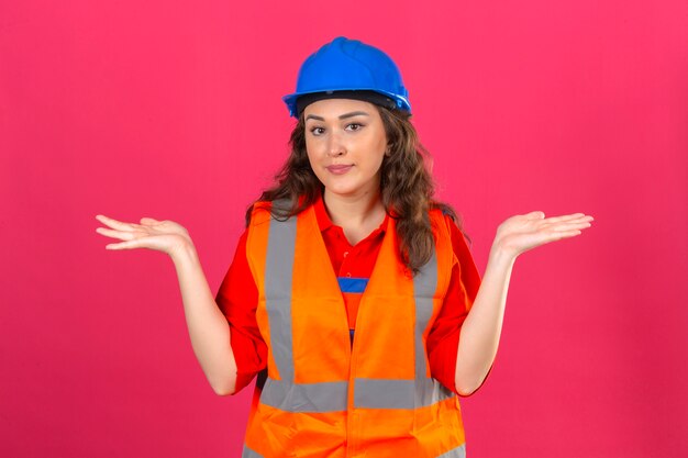 Jeune femme constructeur en uniforme de construction et casque de sécurité désemparé et expression confuse avec les bras et les mains soulevées concept de doute sur mur rose isolé