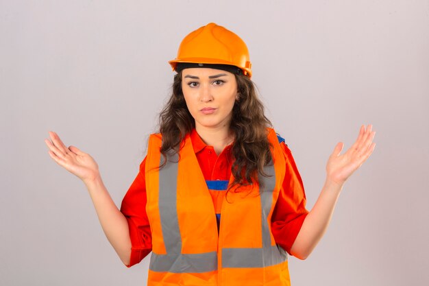 Jeune femme constructeur en uniforme de construction et casque de sécurité expression désemparée et confuse avec les bras et les mains levées sur un mur blanc isolé