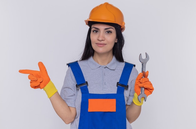 Jeune femme constructeur en uniforme de construction et casque de sécurité en gants de ruber tenant la clé pointant avec l'index sur le côté souriant confiant debout sur mur blanc