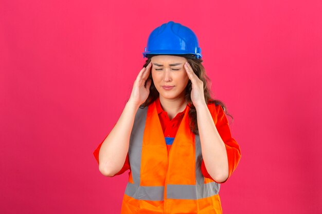 Jeune femme constructeur en uniforme de construction et casque de sécurité à la recherche de mal à toucher sa tête ayant de forts maux de tête sur mur rose isolé