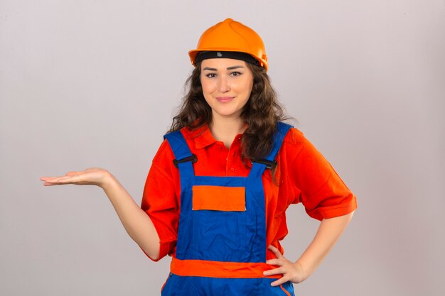 Jeune femme constructeur en uniforme de construction et casque de sécurité souriant gai présentant et pointant avec la paume de la main en regardant la caméra sur un mur blanc isolé