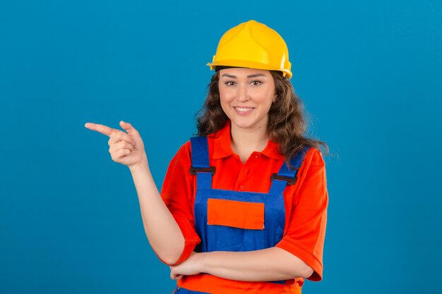 Jeune femme constructeur en uniforme de construction et casque de sécurité souriant joyeusement à la confiance en pointant avec l'index sur le côté sur mur bleu isolé