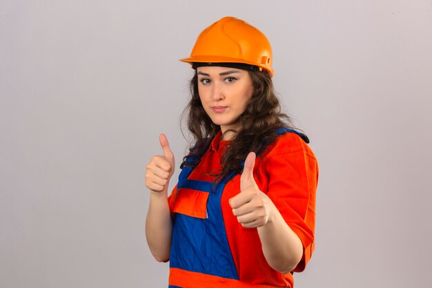 Jeune femme constructeur en uniforme de construction et casque de sécurité avec sourire montrant les pouces vers le haut sur un mur blanc isolé