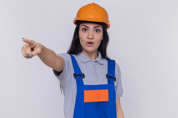 Jeune femme constructeur en uniforme de construction et casque de sécurité à la surprise de pointer avec l'index à quelque chose debout sur un mur blanc