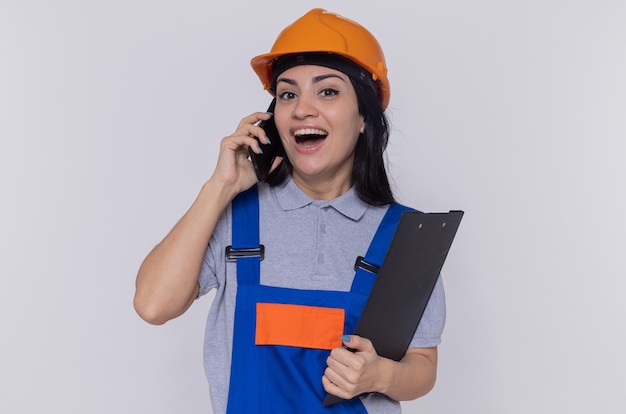 Jeune femme constructeur en uniforme de construction et casque de sécurité tenant le presse-papiers souriant joyeusement tout en parlant au téléphone mobile debout sur un mur blanc