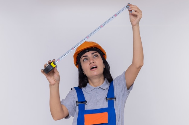 Jeune femme constructeur en uniforme de construction et casque de sécurité tenant un ruban à mesurer en regardant avec un visage sérieux debout sur un mur blanc