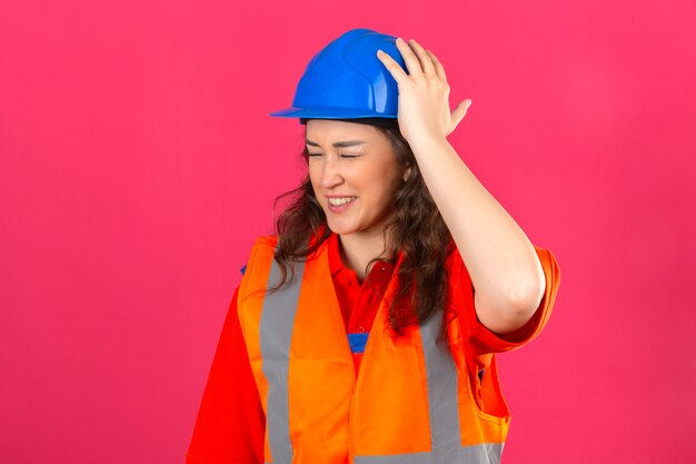 Jeune femme constructeur en uniforme de construction et casque de sécurité à la tête mal en contact avec la douleur sur mur rose isolé