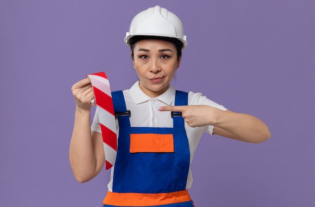 Jeune femme de construction asiatique confiante avec un casque de sécurité blanc tenant et pointant sur une bande d'avertissement