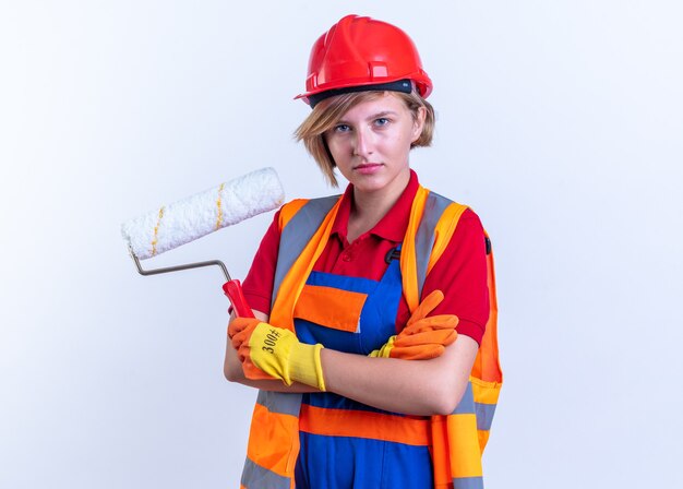 Jeune femme de construction confiante en uniforme avec des gants tenant une brosse à rouleaux traversant les mains isolées sur un mur blanc