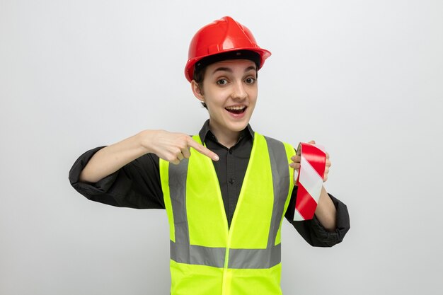 Jeune femme de construction en gilet de construction et casque de sécurité tenant du ruban adhésif pointant avec l'index sur elle souriante et joyeuse