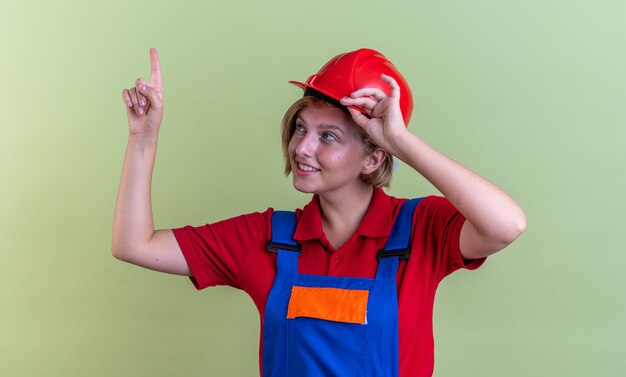 Jeune femme de construction impressionnée en uniforme pointe vers le haut isolé sur un mur vert olive